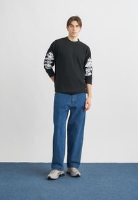 Črni pulover z rebrastim vzorcem in grafičnim tiskom na rokavih, kombiniran z modrimi jeans hlačami. Model nosi sive superge na enobarvnem ozadju.