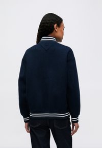 Žena s kučeravými vlasmi, oblečená v námornícky modrom bomberi s bielymi prúžkami na golieri, manžetách a leme, stojaca s chrbtom.