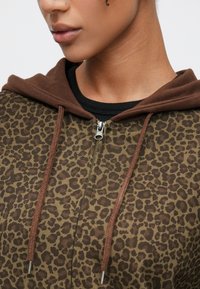 Sudadera con capucha con cremallera y estampado de leopardo marrón, con capucha en marrón a contraste. Cuenta con cordones trenzados y un cuello acanalado negro. Textura de tela suave.