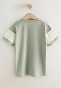 Ljusgrön t-shirt hängande på en trähängare. Har korta ärmar med en kontrasterande crèmefärgad ribbad väv på axlarna och en rund halsringning.