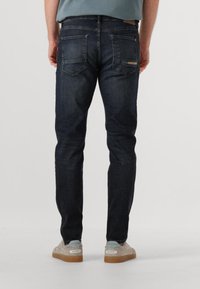 Donkerblauwe spijkerbroek met een slim fit, voorzien van achterzakken en een subtiele logo-accent. Gecombineerd met lichtgekleurde schoenen.