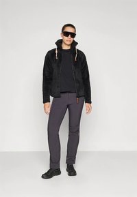 Zwarte fleecevest met een hoge kraag, ritssluiting en verstelbare trekkoorden; gecombineerd met donkergrijze broek en zwart schoeisel.