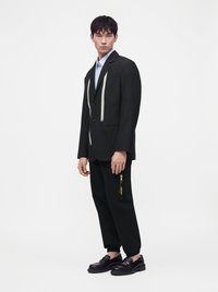 Jeune homme portant un blazer noir avec un ruban à mesurer suspendu, un pantalon noir avec une fermeture éclair latérale, une chemise bleu clair et des mocassins noirs, debout devant un fond blanc.