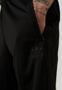 Čierne športové nohavice s vreckom na zips, so subtílnym embosovaným logom Adidas. Hladká, ľahká tkanina s prispôsobeným strihom.