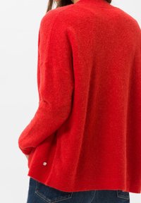 Czerwony sweter z wełny o luźnym kroju, długimi rękawami i małym srebrnym akcentem na dolnym wykończeniu. Miękka, teksturowana powierzchnia.