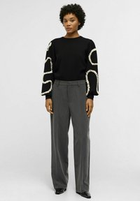 Pull noir avec des bordures décoratives blanches sur les manches, associé à un pantalon gris taille haute. Texture lisse, coupe moderne. Chaussures noires brillantes.