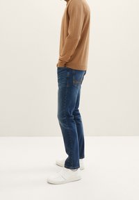 Hombre vistiendo jeans azules, zapatillas blancas y un suéter marrón claro, de pie con las manos en los bolsillos frente a un fondo simple.
