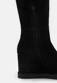 Mustat mokkanahkaiset polven korkeuteen ulottuvat kiilakorkosaappaat, joissa on sivuvetoketju, pyöreä kärki ja teksturoidut korot. Pehmeä materiaali ja tyylikäs siluetti.