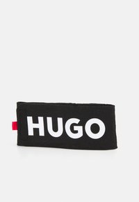 Accesorio de tela rectangular negro con una textura suave, que presenta letras blancas en negrita que dicen "HUGO" en la parte frontal y una etiqueta roja en el lateral.