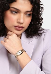 Zlato obarvan ročni ura z pravokotnim številčnicom, belim ciferblatom in kovinskim trakom; nošena na rahlo obarvanem zapestju, v kombinaciji z sivim jopičem.