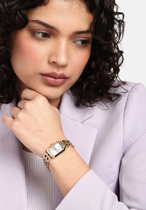 Zlato obarvan ročni ura z pravokotnim številčnicom, belim ciferblatom in kovinskim trakom; nošena na rahlo obarvanem zapestju, v kombinaciji z sivim jopičem.