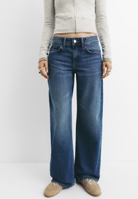 Široke modre jeans hlače z srednjim pasom, rahlo zbledelimi barvami in klasičnim petimi žepi. V kombinaciji z svetlo sivo prilegajočo se srajco.