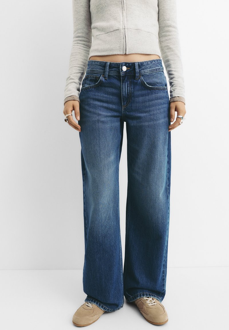 Široke modre jeans hlače z srednjim pasom, rahlo zbledelimi barvami in klasičnim petimi žepi. V kombinaciji z svetlo sivo prilegajočo se srajco.