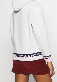Sudadera gris claro con bolsillo tipo canguro, con texto en azul marino y rosa en el dobladillo. Combinada con una falda de pana burdeos.