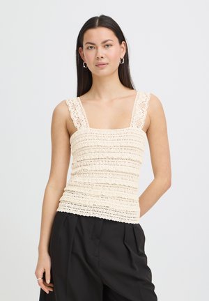 Femme aux longs cheveux noirs portant un haut sans manches en crochet crème et un pantalon noir plissé, debout devant un fond uni.