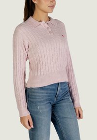 Maglione lavorato a maglia di colore rosa chiaro con motivo a trecce, maniche lunghe e colletto con tre bottoni. Orlo e polsini a coste, abbinato a jeans blu.