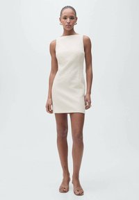 Robe crème courte sans manches avec un design structuré et un tissu texturé, associée à des sandales à glissière nude. Forme simple, accents minimalistes.