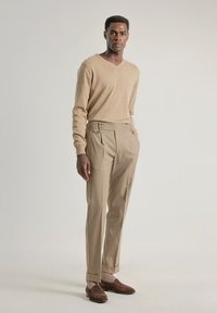 Suéter de punto beige con escote en V, combinado con pantalones claros beige de corte entallado que presentan un detalle de doble botón y puños enrollados. Zapatos de ante marrones.