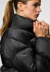 Chaqueta acolchada negra con collar alto, que presenta secciones acolchadas y una textura suave. La tela tiene un brillo sutil y es ligera.
