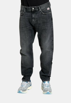 Jeans neri sbiaditi a gamba dritta indossati con scarpe da corsa bianche e blu da una persona su uno sfondo uniforme.