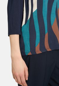 Flerfärgad topp med vågiga mönster i nyanser av blått, elfenben och brunt. Tillverkad av mjukt tyg med tre fjärdedels ärmar.