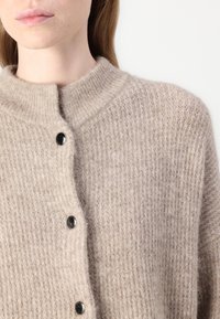 Beige kötött kardigán, texturált mintával, kerek gallérral és fekete gombokkal, lágy, kényelmes megjelenést és érzetet kölcsönöz.