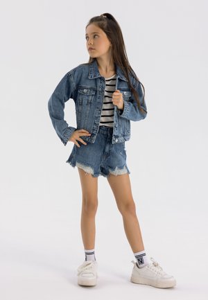 Denim jacket with blue wash, two front pockets, paired with striped shirt, distressed blue denim shorts, and white sneakers with textured soles.