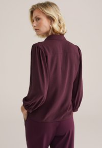 Blouse bordeaux avec des manches ballon et un col, en tissu texturé et à coupe décontractée. Le dos présente un détail de empiècement pour un design supplémentaire.