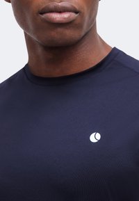 Κοντινό πλάνο ενός ατόμου που φοράει μπλε σκούρο πουκάμισο με μικρό λευκό κυκλικό λογότυπο στο στήθος, εμφανίζοντας το κάτω μέρος του προσώπου και τον λαιμό.