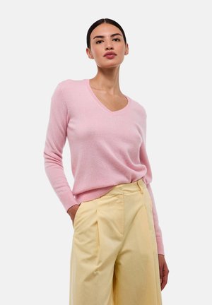 Femme portant un pull V à col rose rentré dans un pantalon beige taille haute et large, se tenant avec une main dans la poche.