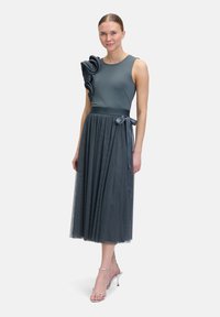Robe grise avec un corsage ajusté, une épaule ornée de volants en tissu superposés, et une jupe fluide en tulle. Ruban en satin à la taille.