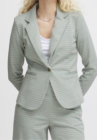 Světle zelený kostkovaný blazer vyrobený z texturované látky; obsahuje špičaté klopy, jeden knoflík a přední kapsy. V kombinaci s bílým topem.