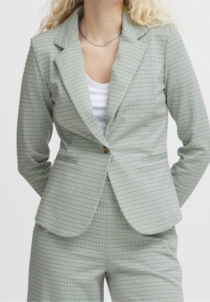 Světle zelený kostkovaný blazer vyrobený z texturované látky; obsahuje špičaté klopy, jeden knoflík a přední kapsy. V kombinaci s bílým topem.