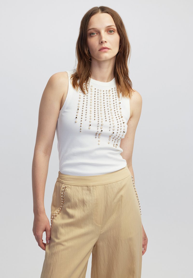 Camiseta de tirantes blanca con texturizado y cuentas doradas dispuestas en líneas verticales. Combinada con pantalones beige que tienen un detalle de cuentas en los laterales.