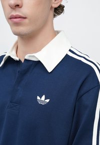 Kék galléros póló fehér bordázott gallérral, három fehér csíkkal az ujjakon és egy kis fehér Adidas logóval a mellkason.