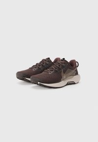 Baskets de sport marron avec une tige en mesh respirant, un orteil renforcé et une semelle beige contrastante, dotées d'accents texturés et de lacets.