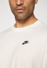 Mand med lys ansigtshår iført en ensfarvet off-white Nike t-shirt med et lille sort logo på venstre bryst.