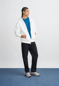 Biela mikina na zips s modrým tričkom, čierne nohavice a šedé tenisky. Model stojí na modrom koberci, s rukami v vreckách.
