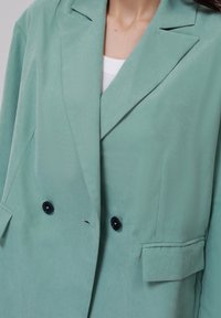 Mintgrønn blazer med en glatt tekstur, som har et dobbeltbrettet design, sorte knapper og to frontlommer.