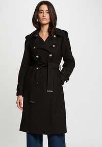 Cappotto lungo nero con design doppiopetto, bottoni argentati, ampi rever e vita con cintura. Presenta maniche lunghe e un taglio al ginocchio.