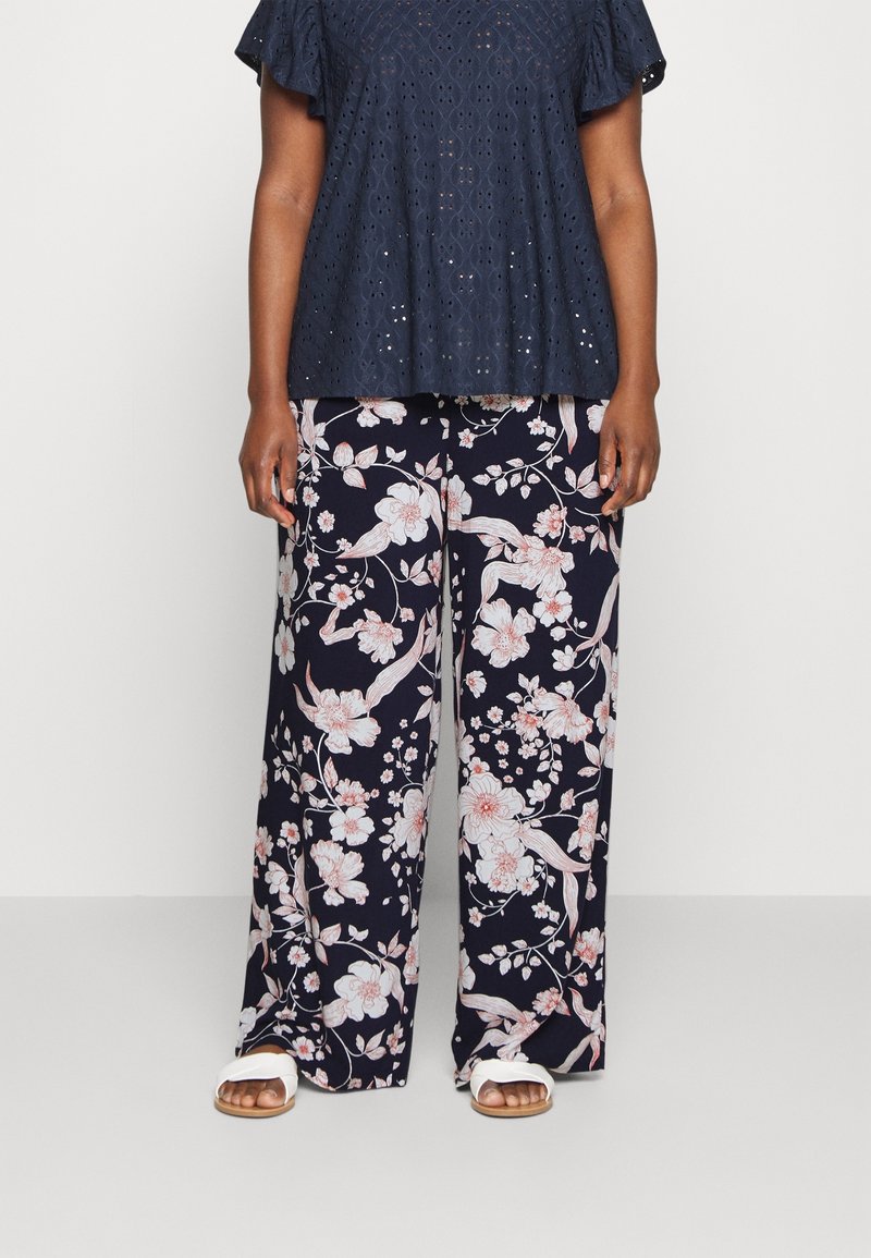 Maritiem bloemen top met oogjespatroon, gecombineerd met wijde broek met grote roze en witte bloemenpatronen. Witte sandalen maken de outfit compleet.