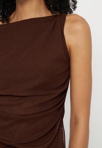Primer plano de una camiseta sin mangas marrón con detalle fruncido en el lateral, llevada por una persona con cabello rizado y tono de piel medio.