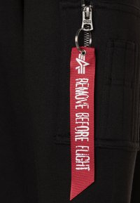 Crne tkanine joggeri imaju zatvarač s metalnom ručkom i crvenu oznaku koja glasi "Ukloniti prije leta" u tamnobelim slovima.