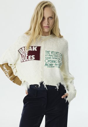 Weißer, kurz geschnittener Pullover mit strukturiertem Strick, der grafische Aufnäher in Bordeauxrot und Grün aufweist. Akzentuiert mit braunen Ärmel-Details und fransigen Kanten.
