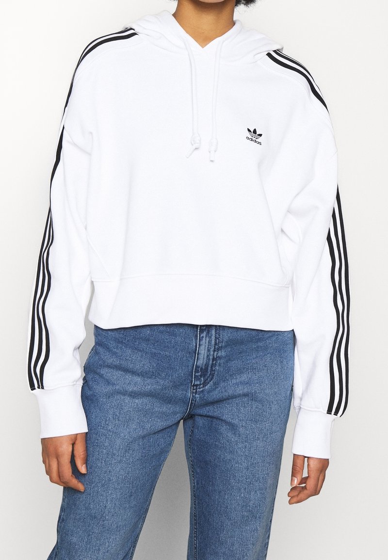 Haut à capuche blanc coupé avec un design à trois rayures noires sur les manches et un petit logo noir sur la poitrine, fabriqué en tissu doux avec un cordon de serrage.