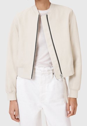 Personne portant une veste bomber beige clair avec une fermeture éclair ouverte sur un t-shirt blanc et un pantalon blanc taille haute avec une ceinture effilochée.
