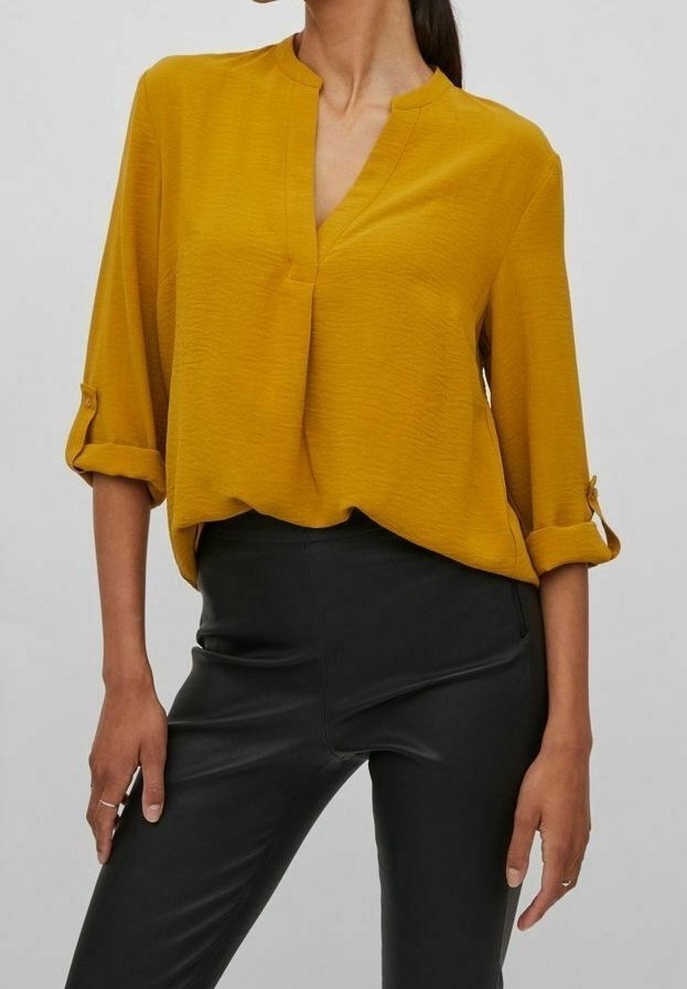 Femme portant un chemisier jaune moutarde à manches longues avec les poignets retroussés et un pantalon noir ajusté, vue des épaules aux hanches.