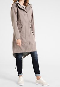 Cappotto impermeabile grigio chiaro con cappuccio, chiusura frontale con cerniera e vita regolabile. Presenta un orlo posteriore più lungo e tasche laterali con zip. Indossato sopra jeans e sneakers.