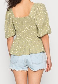 Grøn bluse med polka prikker, puffede ærmer og smocket talje, har et firkantet rygdesign. Kombineret med lyseblå slidte denimshorts.