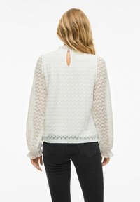 Blouse blanche avec un dos texturé et des manches transparentes à motifs, présentant un détail en trou de serrure au niveau du cou et des poignets volants. Associée à un jean noir.